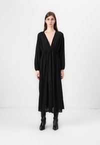 Μαύρο μακρύ φόρεμα με βαθύ V-λαιμό, συγκεντρωμένο στη μέση, με μακριά μανίκια και υφή στο ύφασμα. Φοράει μαύρες μπότες μέχρι το γόνατο.