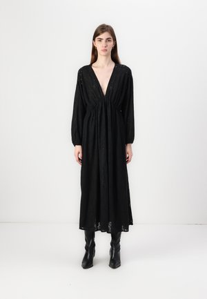 Abito lungo nero con scollatura a V profonda, arricciato in vita, maniche lunghe e tessuto texturizzato. Indossato con stivali neri fino al ginocchio.