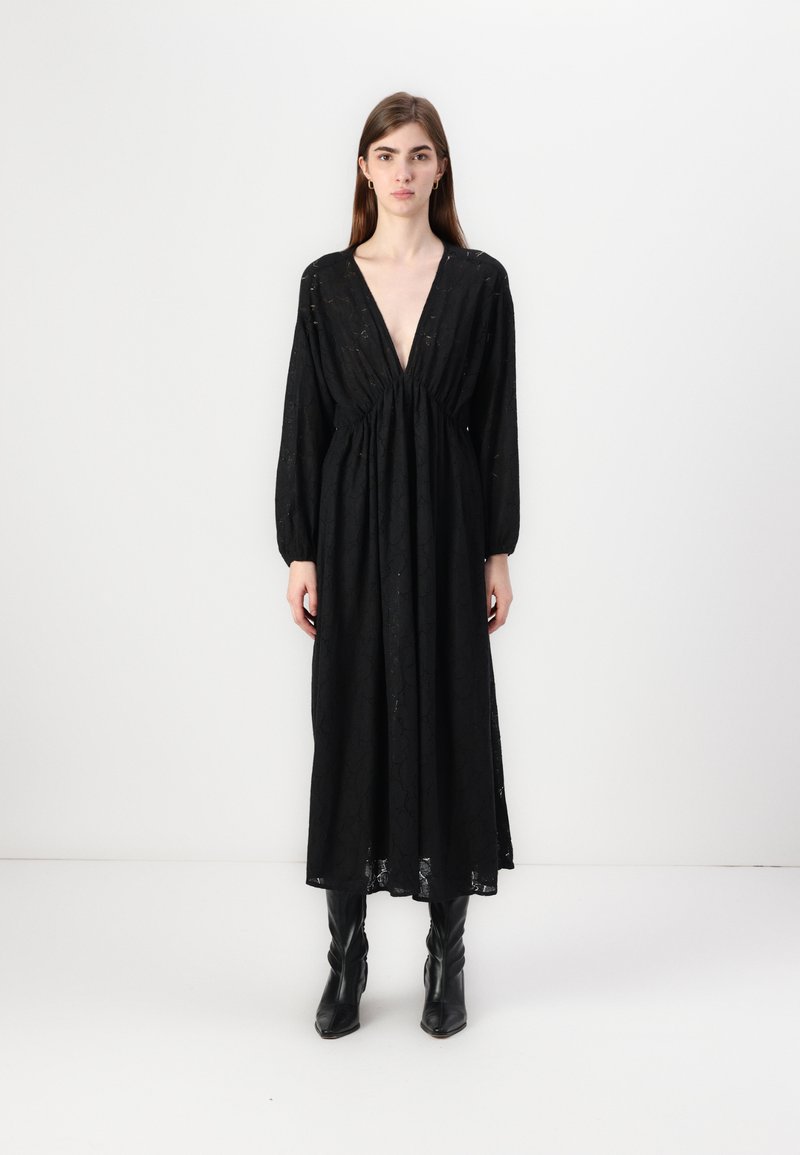 Μαύρο μακρύ φόρεμα με βαθύ V-λαιμό, συγκ�εντρωμένο στη μέση, με μακριά μανίκια και υφή στο ύφασμα. Φοράει μαύρες μπότες μέχρι το γόνατο.