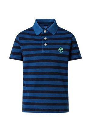 Garçon debout les mains dans les poches, portant un polo à rayures bleues, un pantalon cargo beige et des baskets blanches sur un fond uni.