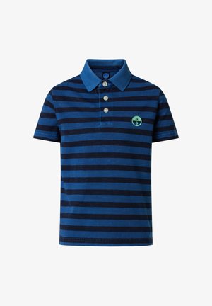 Garçon debout les mains dans les poches, portant un polo à rayures bleues, un pantalon cargo beige et des baskets blanches sur un fond uni.