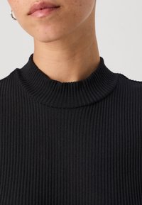 Črn zgibani majica z lahnim ovratnikom in oprijetim krojem, ki prikazuje teksturirano površino in visok ovratnik. Zasnovana za udobno nošenje.