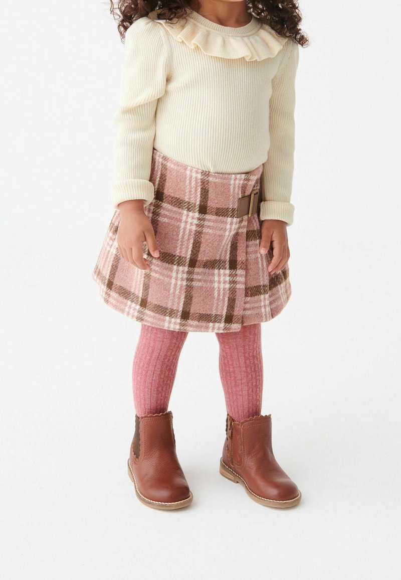 Enfant portant un pull crème à volants, une jupe rose à carreaux avec boucle, des collants roses et des bottines marron, debout sur fond blanc.
