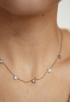 Zarte Silberkette mit kleinen blumenförmigen Gliedern und zentralen Steinen, die auf einem nackten Hals ruht, vor einem neutralen Hintergrund.