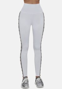 Białe legginsy z wysokim stanem, czarnymi paskami po bokach z akcentami i gładką fakturą. W połączeniu z wzorzystymi butami sportowymi.