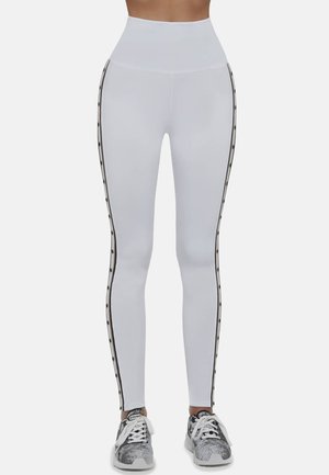 Białe legginsy z wysokim stanem, czarnymi paskami po bokach z akcentami i gładką fakturą. W połączeniu z wzorzystymi butami sportowymi.