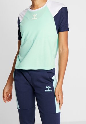 Person trägt Sportkleidung von hummel in Minzgrün, Weiß und Marineblau mit Logos auf Brust und Oberschenkel vor einfarbigem Hintergrund.