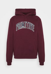 Sweat à capuche bordeaux avec poche kangourou à l'avant et texte "PRIMITIVE" en noir et blanc, incurvé sur la poitrine.