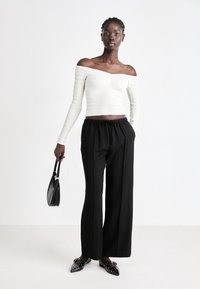 Bílý žebrovaný top bez ramínek, černé volné kalhoty, černá kabelka, špičaté baleríny s bílými cvočky. Jednoduché pozadí, neutrální barvy.