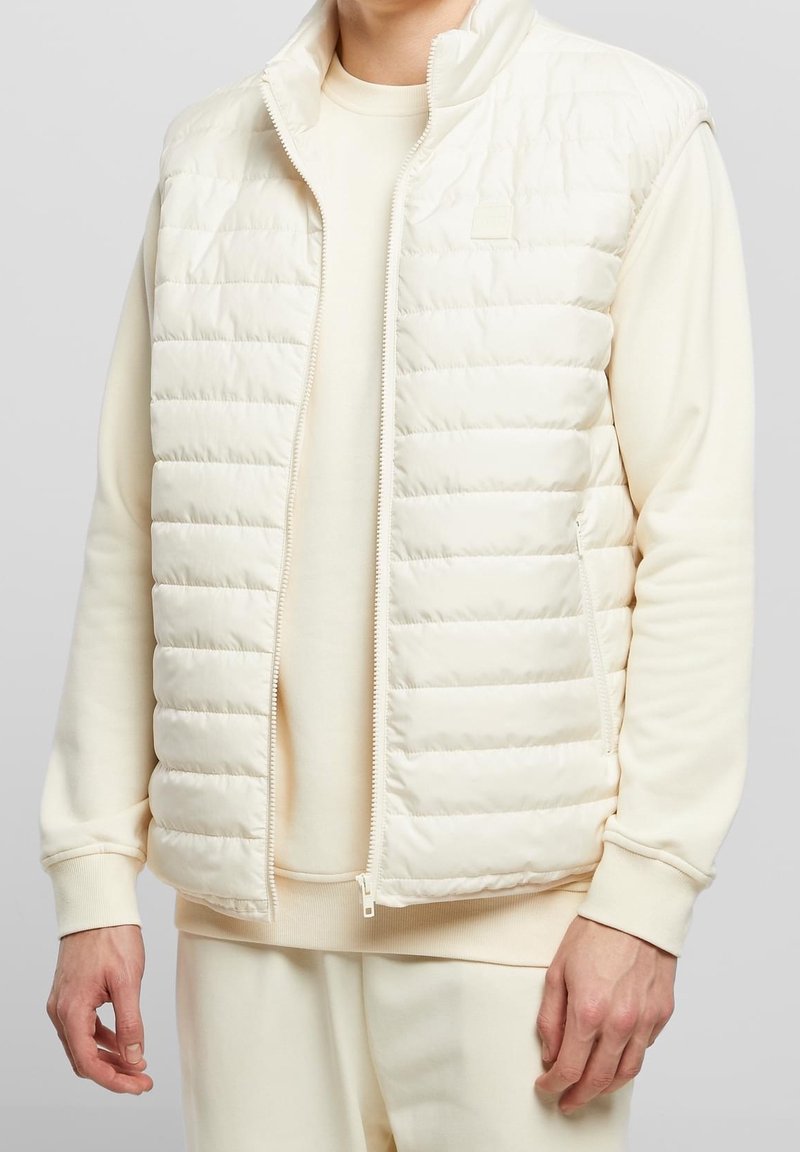 Veste sans manches matelassée couleur crème clair avec texture matelassée, col montant et fermeture éclair à l'avant. Portée sur un sweat-shirt crème assorti, mettant en valeur un design épuré.