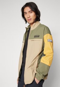 Giacca leggera a blocchi di colore con torso beige, maniche verdi e dettagli gialli. Presenta tasche con zip e polsini elasticizzati.