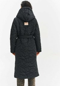 Žena nosi dlhý čierny prešívaný kabát s kapucňou a opaskom, stojí chrbtom kúsok chrbta, pod kabátom sú viditeľné nohavice s leopardím vzorom.