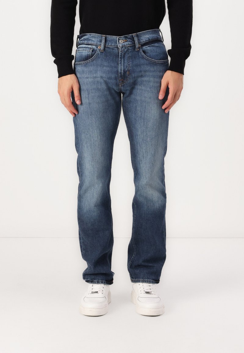 Jean en denim bleu avec une coupe droite, cinq poches et un lavage délavé. Dispose d'une fermeture à bouton et à fermeture éclair. Porté avec des baskets blanches.