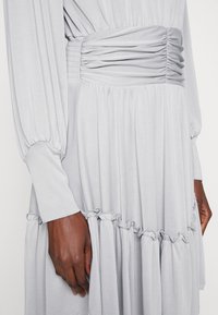 Robe à manches longues gris clair avec taille froncée et volants superposés, portée par une personne à la peau foncée.