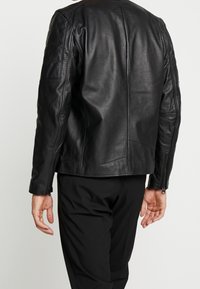 Giacca in pelle nera con una textures liscia, design aderente e maniche con zip. Presenta un colletto alto e nessun logo visibile.