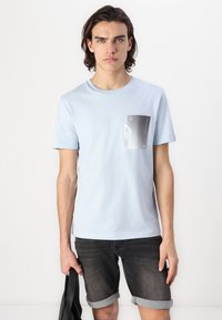 T-shirt en coton bleu clair avec un imprimé graphique noir et blanc sur la poitrine gauche. Manches courtes, décolleté rond, coupe décontractée.