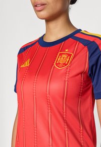 Woman wearing a red and navy Spain soccer jersey with yellow stripes and the Spanish Football Federation crest on chest.