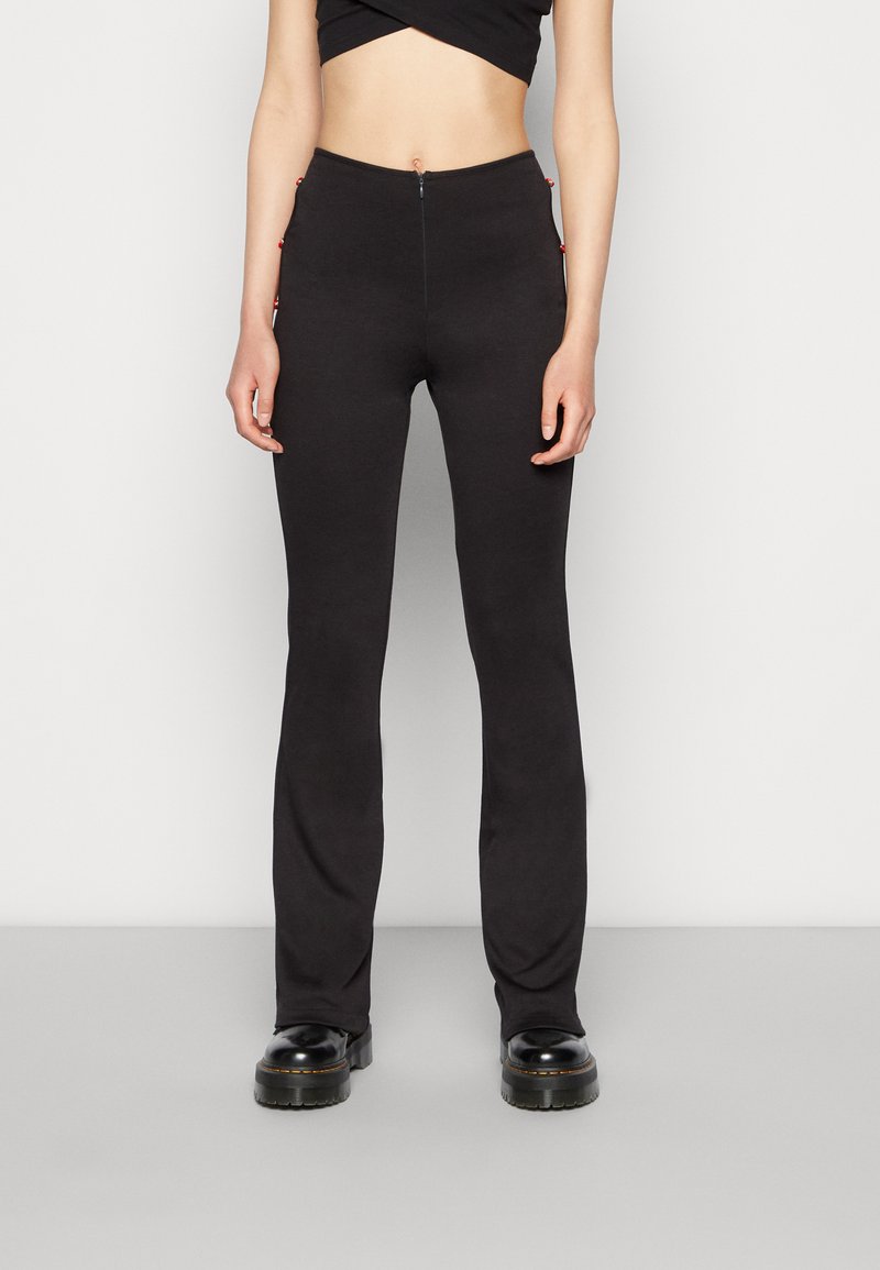 As calças pretas com boca de sino apresentam uma textura suave, cintura alta e detalhes vermelhos nos bolsos laterais. Projetadas para uma silhueta ajustada.