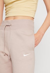 Ljusbeige svettbyxor med dragsko i midjan och vitt Nike-logotyp, bärs med en gul crop top, hand i fickan synlig.