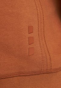 Detail zblízka na rezavě zbarvenou látku s třemi malými vertikálními geometrickými gumovými nášivkami a viditelnými detaily švů.