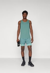Tank top and shorts in teal, featuring a breathable fabric. Model holds a black water bottle, wearing black sneakers and socks.