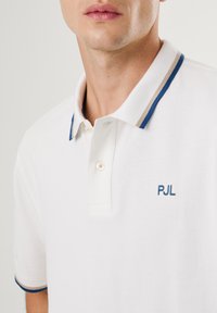 Biely polo tričko s textúrovanou tkaninou, kontrastne modro-beige pruhovaným golierom a vyšitým logom "PJL" v modrej farbe na hrudi.