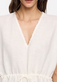 Witte linnen kledingstuk heeft een diepe V-halslijn met rauwe randen, een geraffineerde taille met een gestrikt koord en een relaxed fit.
