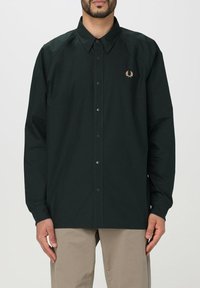 Camicia a maniche lunghe verde scuro con colletto a punta, dotata di un piccolo logo ricamato sul petto e bottoni a pressione lungo la parte anteriore.