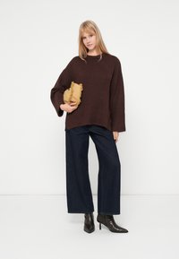 Καφέ πλεκτός σκούφος με φαρδιά μανίκια, σε συνδυασμό με σκούρα μπλε φαρδιά παντελόνια και μαύρες ψηλοτάκουνες μπότες με尖尖. Κρατάει μια χρυσή τσάντα με φούντες.