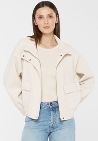 Woman with straight brown hair wearing a cream zip-up jacket over a light top and blue high-waisted jeans, standing with hands on hips.