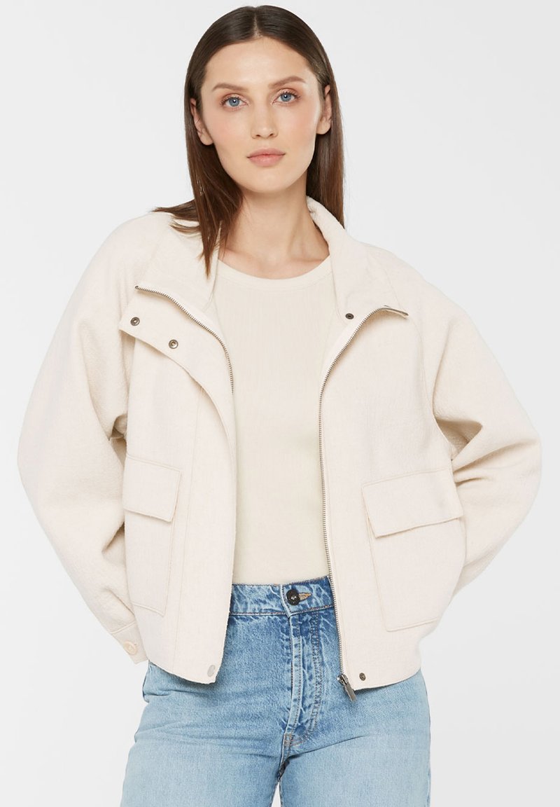 Woman with straight brown hair wearing a cream zip-up jacket over a light top and blue high-waisted jeans, standing with hands on hips.