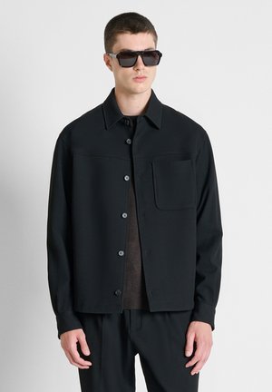 Jonge man die een zwart jasje met knopen en borstzak draagt, donkere zonnebril en donkere broek, staand tegen een effen lichte achtergrond.