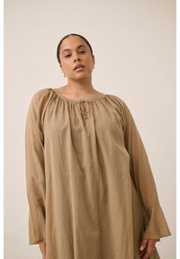 Femme aux cheveux tirés en arrière, portant un chemisier beige ample à manches longues avec un col froncé et un détail à nouer, debout devant un fond uni.