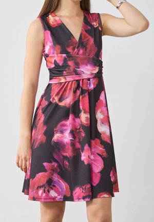 Sukienka bez rękawów z dekoltem w serek, czarna z dużym, żywym różowo-czerwonym kwiatowym wzorem, noszona przez kobietę z uniesioną ręką i bransoletką.