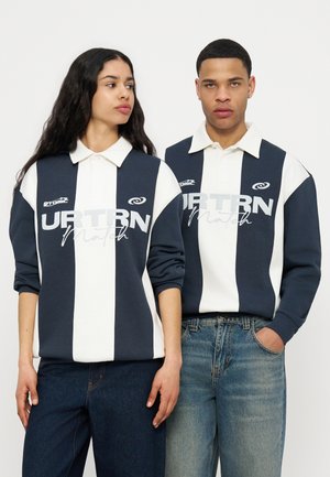 Twee mensen dragen bijpassende marineblauwe en witte rugbyshirts met strepen en de tekst "URTRN Match"; de ene persoon kijkt opzij, de andere kijkt vooruit.