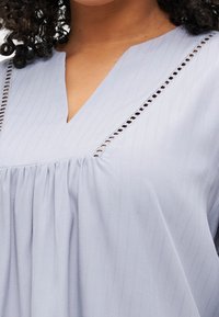 Haut bleu clair avec un décolleté en V et des rayures verticales, présentant une texture froncée et des détails transparents le long de l'encolure et des épaules.