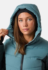 Türkise isolierte Jacke mit einem Reißverschluss, puffiger Gestaltung und Kapuze mit Kordelzug, getragen mit einem schwarzen gemusterten Haarband.