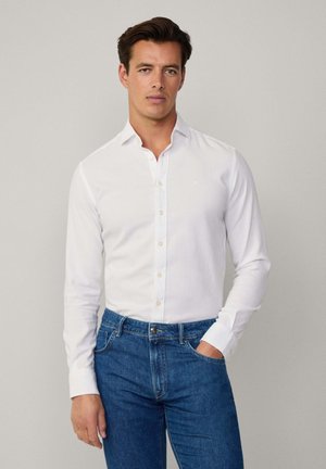 Camisa blanca de manga larga con cuello abotonado, de corte ajustado, combinada con jeans de mezclilla azules. Logo sutil en el pecho de la camisa.