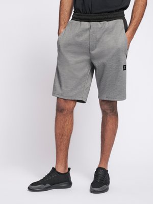 Mand iført grå knælange shorts med sort elastisk linning og sorte sneakers, stående med hænderne i lommerne.