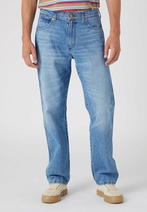Homme portant un jean droit bleu clair, une chemise rayée et des baskets beiges, debout sur un fond blanc uni.
