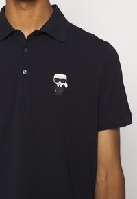 Homme portant un polo noir avec une petite figure dessinée d'une personne aux cheveux blancs et portant des lunettes de soleil sur le côté gauche de la poitrine.