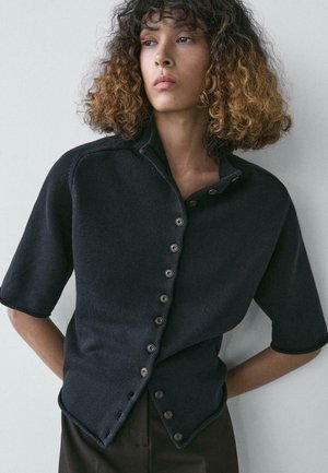 Frau mit lockigem Haar, die eine dunkle, kurzärmelige, geknöpfte Strickjacke trägt und mit verschränkten Händen hinter dem Rücken vor einem hellen, neutralen Hintergrund steht.