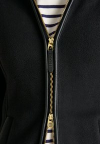 Nahaufnahme einer schwarzen Fleecejacke mit teilweise offenem goldenen Reißverschluss, die ein weißes und marineblau gestreiftes Hemd darunter zeigt.