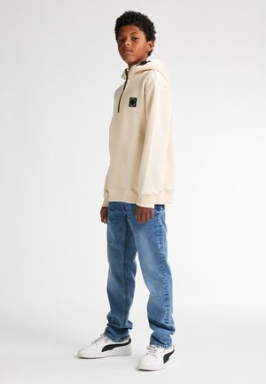 Sweat à capuche beige avec un logo noir sur la poitrine, associé à un jean bleu clair et des baskets blanches avec des accents noirs. Texture de tissu lisse.