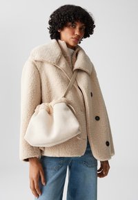Donna con capelli ricci che indossa un cappotto beige a trama e jeans blu, mentre porta una borsa a spalla morbida color beige su uno sfondo grigio uniforme.