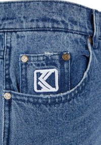 Jeans in denim blu con una tasca posteriore dotata di un logo ricamato bianco, rivetti metallici e una texture invecchiata sul bordo della tasca.