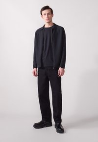 Veste zippée noire sur un t-shirt noir, associée à un pantalon noir et des bottes noires à semelle épaisse, avec un tissu texturé et une silhouette classique.