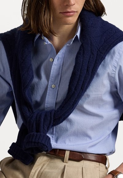 Homem a usar uma camisa de botão azul clara com um suéter azul-marinho pendurado nos ombros, calças bege e um cinto de couro castanho.