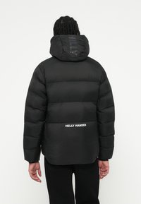Personne avec des cheveux tressés portant une veste matelassée noire avec le logo "Helly Hansen" dans le bas du dos, se tenant devant un fond blanc uni.