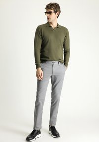 Polo a maniche lunghe verde oliva, pantaloni slim-fit grigio chiaro e sneakers nere con suola bianca, in piedi davanti a uno sfondo neutro.